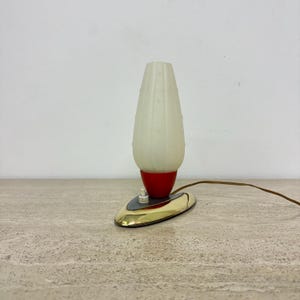 Op de afbeelding: Een witte, gematteerde glazen tafellamp met een rode voet en een gouden metalen voet. De lamp heeft een snoer en een schakelaar.