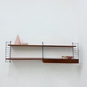 Op de afbeelding: Mid-century modern wandplank met twee planken en een lade. De planken zijn gemaakt van donker hout en worden ondersteund door zwarte metalen draadframes. Een roze kegel en kom staan op de bovenste plank.