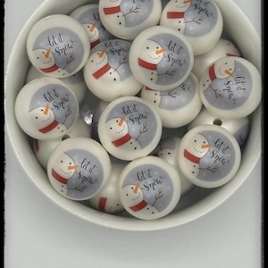 May include: A collection of white, round beads with a snowman design and the words "Let it Snow" in a cursive font. Each snowman has a red scarf and a gray background. The beads are in a white bowl.
