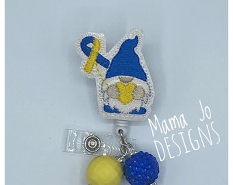 May include: A blue and yellow gnome with a yellow hat and a blue beard. The gnome is holding a yellow ribbon. The gnome is attached to a retractable badge holder with a blue and yellow bead. The badge holder has the text "Mama Jo DESIGNS" on it.