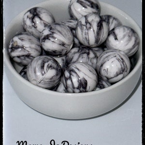 May include: A white bowl filled with black and white marbled round beads. The bowl is sitting on a light blue surface. The text "Mama Jo Designs" is written in black at the bottom of the image.
