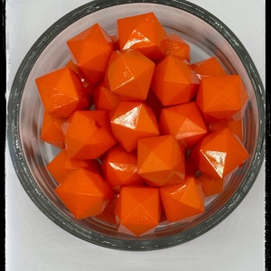 May include: A close-up of a glass bowl filled with bright orange, faceted, geometric beads. The beads are all the same size and shape, and they are arranged in a random pattern. The bowl is sitting on a white surface.