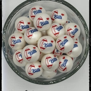 May include: A bowl of white beads with a football and heart design. Each bead has the word "Bills" printed in blue on it. The beads are in a clear glass bowl. The text "Buffalo Beads" is at the top of the image and "Mama Jo Designs" is at the bottom.