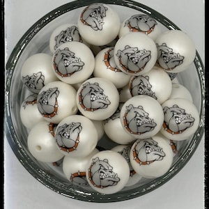 May include: A bowl of white, round beads with a bulldog graphic printed on each bead. The bulldog is gray with a black nose and orange collar.