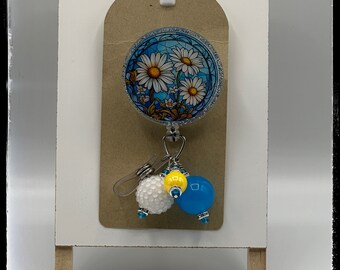 May include: A badge reel with a daisy design, featuring white daisies on a blue background. The reel has a silver glitter border and is attached to a brown tag. It includes a white, yellow, and blue charm. The text "Daisy Badge Reel" and "Mama Jo Designs" are visible.