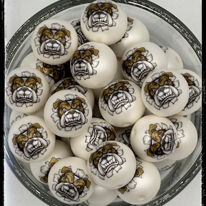 May include: A clear glass bowl filled with white round beads. Each bead features a detailed illustration of a bulldog's face in brown and black, emerging from a torn paper effect. The beads are smooth and have a glossy finish.