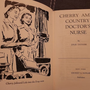May include: Black and white illustration of two nurses in a hospital room. The nurses are standing next to an X-ray machine. The text "Cherry followed Lola into the X-ray room" is at the bottom of the image. The book cover title is "Cherry Ames Country Doctor's Nurse" by Julie Tatham.