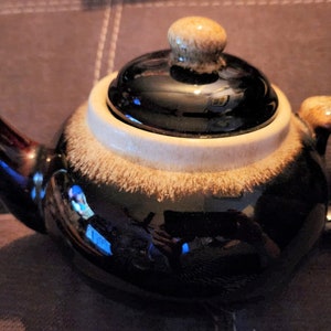 May include: A brown and white ceramic teapot with a lid. The teapot has a speckled glaze and a white band around the top.