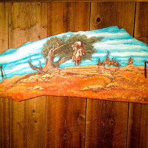 May include: A painting on a wooden panel depicting a Native American on horseback riding through a landscape with a lone tree. The background is a blue sky with white clouds. The painting is framed by a wooden border.