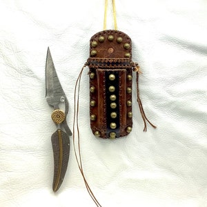 May include: A small, silver knife with a decorative handle and a leather sheath with brass studs. The sheath has a fringe and is hanging from a string.