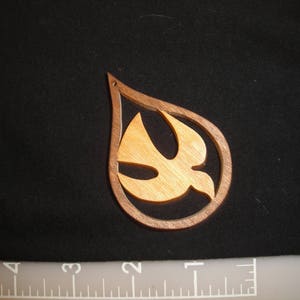 May include: A wooden ornament shaped like a teardrop with a cutout of a dove inside. The dove is made of a lighter wood and is flying upwards.