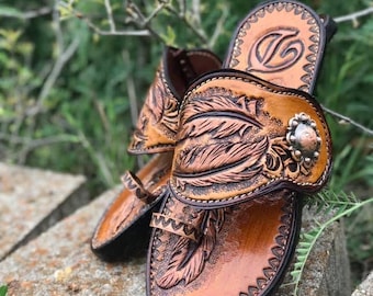 May include: A pair of brown leather sandals with a feather design. The sandals have a strap that goes around the ankle and a toe strap. The sandals are decorated with a silver buckle and a silver bead.