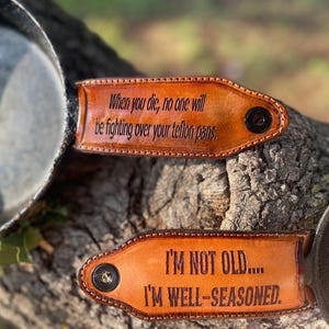 May include: Two brown leather pan handle covers with black rivets are shown. One reads "When you die, no one will be fighting over your teflon pans." The other says "I'M NOT OLD.... I'M WELL-SEASONED."