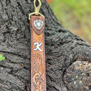 May include: A brown leather keychain with a gold-colored clasp. The keychain features a decorative floral pattern, a silver letter "K", and a silver heart-shaped embellishment. The leather has a distressed appearance.