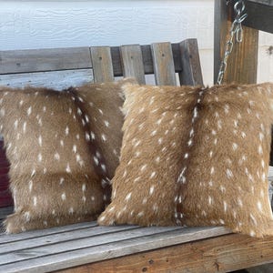 May include: Two square throw pillows covered in brown faux fur with a white spotted pattern and a dark brown vertical stripe running down the center of each. The textured material mimics the appearance of deer hide with soft, dense fibers.