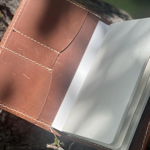 May include: A brown leather notebook cover featuring white contrast stitching, two interior card slots, and a vertical pocket. The cover holds a white paper notebook with rounded corners, showcasing a smooth texture and visible natural leather grain.