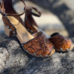 May include: A pair of brown leather clogs with a floral embossed design. The clogs have a wooden sole and a strap around the ankle. Silver studs are placed around the edges of the leather. The clogs are on a textured, gray surface.