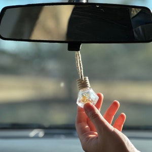 May include: A clear glass car air freshener with a gold-colored liquid inside. The air freshener is hanging from the rearview mirror of a car.