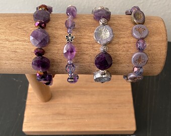 May include: Four purple and silver bracelets with various charms, including a flower, a bee, and a geometric design, displayed on a wooden stand.