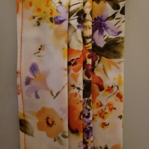 May include: A white face mask with a floral print featuring orange, yellow, and purple flowers. The mask has a pleated design and orange stitching around the edges.