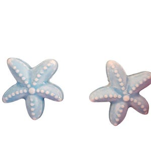 May include: Two light blue starfish-shaped decorative objects. Each starfish has five points and is adorned with small white dots. The objects have a glossy finish and are set against a plain white background.