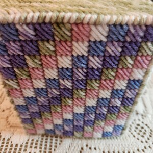 May include: A square tissue box cover with a green, white, pink, and purple plaid pattern. The cover is made of yarn and has a square opening on the top for tissues.
