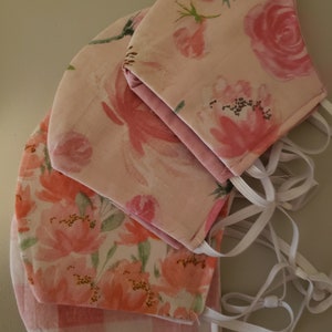 May include: Four pink fabric face masks with floral patterns. The masks are stacked on top of each other, with the top mask having a larger floral pattern and the bottom mask having a smaller floral pattern. The masks have white elastic straps.