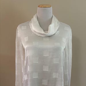 May include: A white blouse with a cowl neck and long sleeves. The fabric has a subtle square pattern. The blouse is made of a lightweight, silky material. The cuffs are finished with a delicate trim.