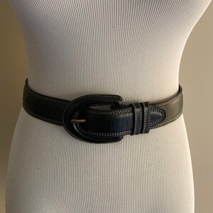 May include: A black leather belt with a large oval buckle and white stitching. The buckle has a gold-toned finish.