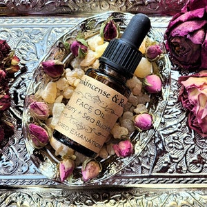 May include: A small amber glass bottle with a black dropper top, filled with a clear liquid. The bottle is labeled "FACE OIL" with the text "Frankincense & Rose" and "Berry + Sea Buckthorn" on the label. The bottle is sitting on a silver tray with dried rose buds and amber-colored crystals.
