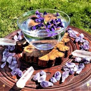 May include: A clear glass bowl filled with water and floating purple flowers sits on a wooden carved tray. Amethyst crystals and clear quartz crystals surround the bowl, creating a spiritual or decorative display.