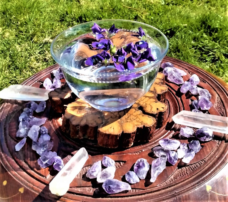 May include: A clear glass bowl filled with water and floating purple flowers sits on a wooden carved tray. Amethyst crystals and clear quartz crystals surround the bowl, creating a spiritual or decorative display.
