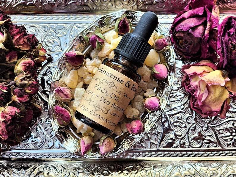 May include: A small amber glass bottle with a black dropper top, filled with a clear liquid. The bottle is labeled "FACE OIL" with the text "Frankincense & Rose" and "Berry + Sea Buckthorn" on the label. The bottle is sitting on a silver tray with dried rose buds and amber-colored crystals.