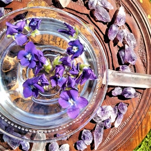 May include: A clear glass bowl filled with water and floating purple flowers, set on a carved wooden tray. Amethyst crystals and clear quartz points surround the bowl, creating a natural, spiritual aesthetic.