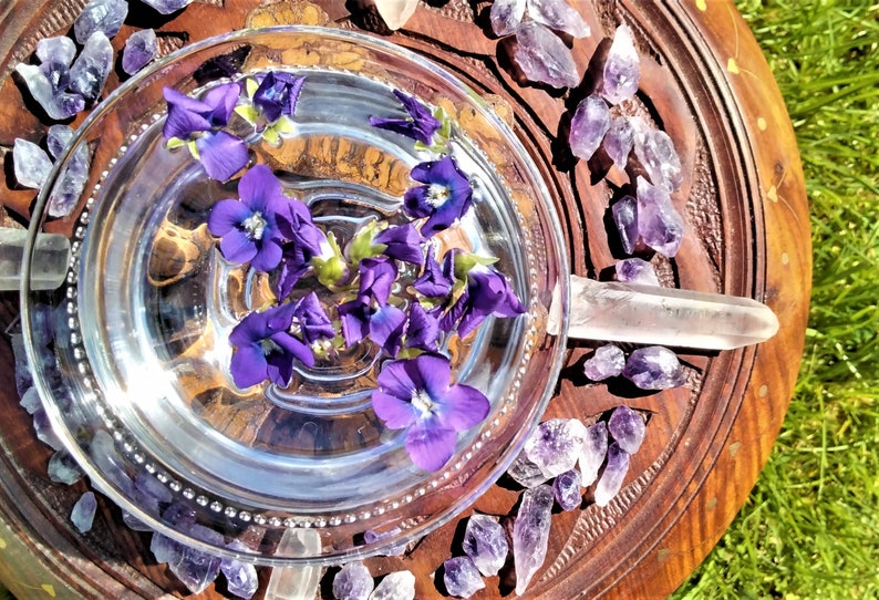 May include: A clear glass bowl filled with water and floating purple flowers, set on a carved wooden tray. Amethyst crystals and clear quartz points surround the bowl, creating a natural, spiritual aesthetic.