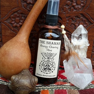 May include: A brown glass bottle with a black spray top and a white label that reads "The Shaman Energy Clearing Mist" and "Eden Apothecary". The bottle is surrounded by a wooden surface, a gourd, a crystal, and a feather.