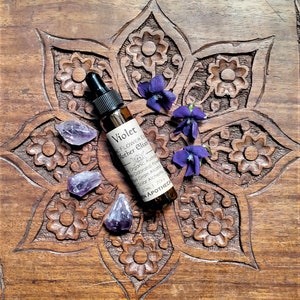 May include: A small brown glass bottle labeled "Violet Flower Essence Mother Elixir" with a black dropper, surrounded by purple amethyst crystals and violet flowers. The bottle rests on a carved wooden surface.