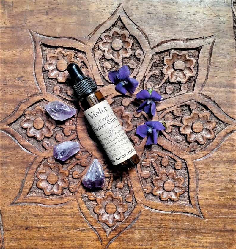 May include: A small brown glass bottle labeled "Violet Flower Essence Mother Elixir" with a black dropper, surrounded by purple amethyst crystals and violet flowers. The bottle rests on a carved wooden surface.