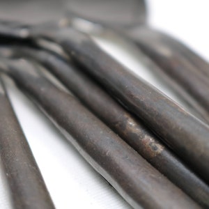 Könnte beinhalten: Ein Set aus sechs schwarzen Löffeln aus Metall mit rustikalem Finish. Die Löffel sind übereinander gestapelt, die Griffe ragen nach rechts heraus.