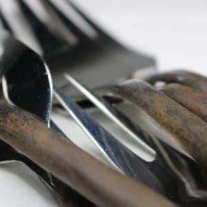 May include: A close-up of a set of silverware, including a fork, a spoon, and a knife. The silverware is made of metal and has a shiny finish. The silverware is arranged in a way that shows off its details.
