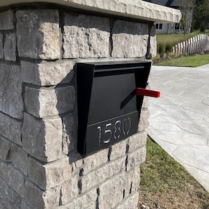 May include: A black mailbox with the number 1580 in a modern font is mounted on a stone pillar. The mailbox has a red flag extended. The pillar is made of gray stone and is topped with a lighter stone.