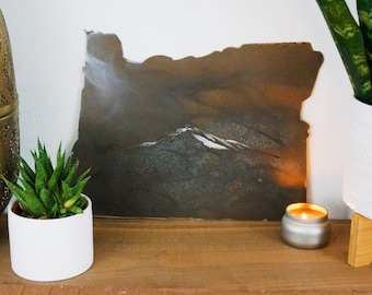 May include: A rusty metal cutout of the state of Oregon with a mountain range silhouette in the center. The cutout is displayed on a wooden shelf with a small potted succulent plant and a silver candle.