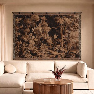 May include: A large rectangular woven wall tapestry featuring a dark brown and beige forest landscape scene with trees and foliage, suspended from a black metal rod above a cream-colored sectional sofa and a round wooden coffee table.