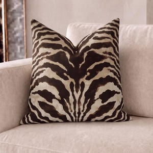 May include: A square throw cushion featuring a dark brown and cream zebra stripe pattern. The fabric displays a symmetrical animal print design with bold, irregular dark stripes set against a lighter, neutral-toned background across the surface.