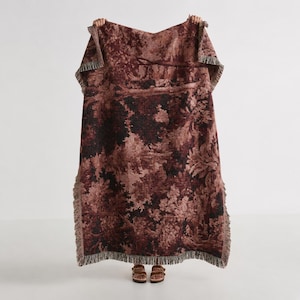 May include: A rectangular woven throw blanket featuring a dark maroon and black toile-style landscape pattern with dense foliage and trees. The blanket has a textured surface and is finished with short, frayed fringe edges along all four sides.