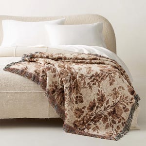 Puede incluir: Una manta con estampado floral beige y marrón con flecos. La manta está sobre una cama con almohadas y sábanas blancas. El estampado floral presenta grandes flores y follaje en tonos marrones.