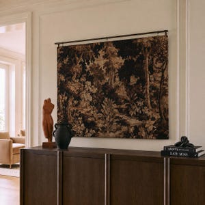 May include: A rectangular wall tapestry featuring a dark, monochromatic forest scene with dense foliage and trees, suspended from a thin metal rod. The tapestry displays intricate, textured patterns in shades of black, brown, and tan.