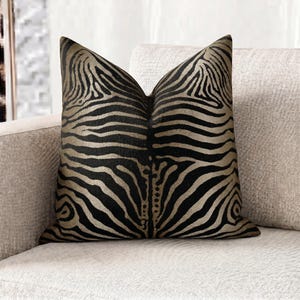 May include: A square cushion featuring a black and gold zebra stripe design. The cushion has a textured surface and is placed on a neutral-toned sofa. This decorative item adds a touch of sophistication to any room. The cushion is a home decor item.