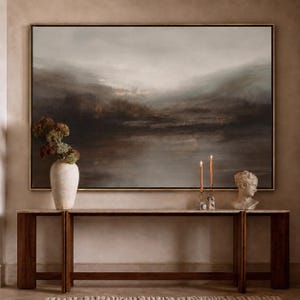 May include: A large, framed abstract landscape painting in shades of gray, brown, and blue hangs above a wooden console table. A white vase with dried flowers, two lit candles, and a bust of a classical figure are on the table.