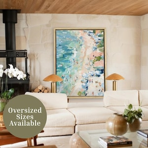 May include: An abstract beach scene painting in a gold frame hangs above a cream-coloured sofa. The artwork features turquoise water, a sandy beach, and figures. Two gold lamps with wooden shades flank the painting. The text "Oversized Sizes Available" is in the foreground.